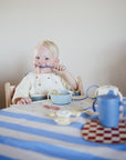 A young child with blonde hair smiles while eating noodles at a table covered with a blue and white striped tablecloth, drinking from the Mushie Trainer Sippy Cup. A teapot and the easy-grip Mushie cup are also on the table.