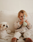A young child in light clothes relaxes on a white couch, sipping from the mushie Silicone Puree Pouch. Their small white dog with tan ears sits beside them—both enjoying a calm, mess-free feeding moment.