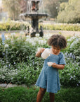 A young girl in a blue dress enjoys imaginative pretend play, pouring water from the Mushie Silicone Tea Play Set into a teacup while standing in a flower-filled garden with a fountain in the background.