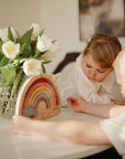 Two children enjoy screen-free play with the mushie Wooden Magnetic Rainbow Maze on a table next to white tulips. The girl watches as the boy interacts with the toy, supporting creativity and fine motor skills in a cozy setting.