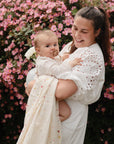 A woman in a white dress smiles while holding a baby with a pacifier before pink flowering bushes. The baby is wrapped snugly in the mushie Organic Cotton Muslin Swaddle Blanket.