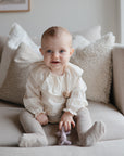 A baby with light hair and blue eyes sits on a beige couch, wearing a cream outfit and holding the mushie Cat Figurine Teether. Decorative pillows are arranged behind them.