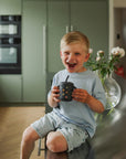 Smiling boy in blue shirt holds a Mushie Silicone Training Cup + Straw on the kitchen counter.