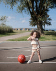 A young child with curly hair poses playfully on an outdoor basketball court beside a red ball, with a mushie Silicone Puree Pouch nearby—ideal for easy, mess-free feeding under the blue sky.