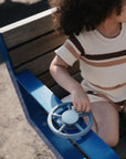 A young child in a striped outfit sits on a wooden bench with blue edges, playing with the mushie Suction Spinner Toy attached to the bench for an engaging sensory experience.