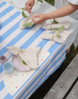 A person arranges green leaf napkins on a blue and white striped tablecloth with pastel plates, cups, and a mushie Silicone Placemat. The outdoor setup is on grass near a wooden bench.