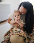 A woman lovingly hugs and kisses her baby, who gazes at the camera while gripping a mushie Unicorn Figurine Teether and is partly wrapped in a vibrant, checkered blanket.