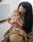 A woman lovingly hugs and kisses her baby, who gazes at the camera while gripping a mushie Unicorn Figurine Teether and is partly wrapped in a vibrant, checkered blanket.