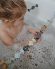 A young child with light brown hair plays in the bath, holding mushie's Daisy Silicone Suction Builders—colorful connected shapes and flowers that help develop motor skills during bath time.