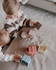 Two children play with the mushie Mix and Match Present Toy, exploring colorful gift boxes. The older child holds a brown box while the younger reaches for a blue one, building fine motor skills with these openable, empty containers.