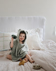 A smiling child sits on a bed wrapped in the mushie Bear Poncho Towel, featuring cute bear ears. Surrounded by small toys, the cozy scene is set in a softly lit room with white bedding and pillows.