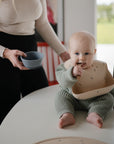 A baby in a green knit outfit sits on a table, wearing a bib and holding a mushie Silicone Toddler Starter Spoon, ready for self-feeding. An adult with a gray bowl stands nearby; greenery is visible through the window.