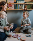 A woman and two children sit on the floor, smiling and playing with the mushie Foam Bath Puzzle. Books on a wall shelf behind them emphasize the educational benefits of hands-on learning together.