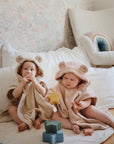 Two babies in bear-ear hooded towels sit on a soft blanket. One holds a snack, while the other plays with the mushie Nesting Stars Toy, great for fine motor skills. Colorful blocks, a white chair, rainbow pillow, and patterned wallpaper complete the scene.
