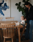 A woman secures a baby in a high chair at a wooden dining table set with mushie Silicone Placemat, baby utensils, toys, and a mug. Wooden chairs, a lush green plant, and abstract art create a cozy background.