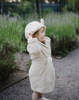 A toddler beams in the mushie Bear Robe—an organic cotton hooded robe with adorable ears—while standing on a garden path. Lush green plants create the perfect backdrop for this cozy baby essential.