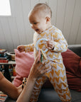 A smiling baby in a yellow heart-patterned onesie stands on a couch, holding the mushie Western Teething Ring, supported by an adult, with colorful pillows and books in the background.