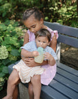 A young girl in a pink dress sits on a gray bench outdoors, holding a baby dressed in stripes. The baby enjoys mess-free feeding from the mushie Silicone Puree Pouch. Green plants and flowers decorate the background.
