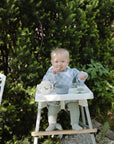 A baby sits outdoors in a high chair, wearing a blue and white checkered bib and holding Mushie First Feeding Baby Spoons, with a bowl and sippy cup on the tray, surrounded by lush green bushes.