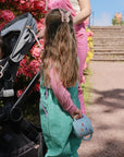 A young girl in green overalls holds a mushie Snack Cup beside a stroller with a toddler. An adult in pink stands nearby outdoors, near blooming pink flowers and stone steps.