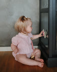 A blonde toddler in a pink outfit sits on a wooden floor, playing with the mushie Suction Spinner Toy by the glass door of a dark cabinet. The child enjoys the sensory play, gazing at their reflection with bottles and glassware inside.