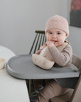 A smiling baby in a pink knit hat and beige outfit sits in a high chair, holding a bib and chewing on fingers, with Mushie First Feeding Baby Spoons and a bowl placed on the table nearby.