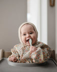 A smiling baby in a floral outfit and knit bonnet sits in a high chair, happily chewing on a Mushie First Feeding Baby Spoon, the softly lit background highlighting this adorable first feeding moment.