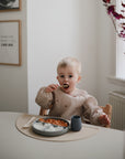 A young child with blonde hair sits at a table, eating from a divided plate and using Mushie First Feeding Baby Spoons. In the background, wall art, a window, and a vase with pink leaves can be seen.