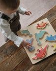 A young child stands at a wooden table, playing with the Mushie Wooden Puzzle, placing colorful dinosaur-shaped pieces onto the board to build fine motor skills.