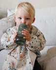 A young child with blonde hair sits on a light-colored couch, wearing a colorful, water resistant mushie Long Sleeve Bib with space-themed patterns and drinking from a green sippy cup.