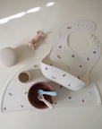 The mushie Silicone Placemat displays a baby feeding set: a patterned bib, pink bowl, beige cup, two spoons, a pink toy, and a ribbed container—all arranged on its food-grade surface with playful colorful prints.