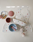 A baby feeding set with a polka dot bib, matching mushie Silicone Placemat, pink and blue bowls with child utensils, a textured pink lid, elephant toy ring, and cream rattle placed on a light background.