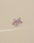 The mushie Cat Figurine Teether, a small purple cat-shaped baby teether with a curled tail, stands on a smooth beige surface under soft lighting.