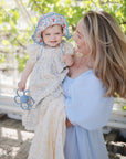 A smiling woman in a light blue dress holds a happy toddler wearing a floral sunhat and dress. The child clutches a blue toy and the mushie Organic Cotton Muslin Swaddle Blanket. Sunlight and greenery fill the background.