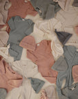 A flat lay of mushie Zipper Pajamas in soft pastel hues—pink, blue, cream, and gray—neatly arranged on a neutral background among baby essentials like onesies, sweaters, and hats.
