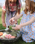 Two young girls pour water into a birdbath with Mushie Whale Bath Stackers on the lawn.
