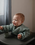 A smiling baby in a high chair uses a Mushie First Feeding Baby Spoon while wearing a light blue bib. A green cup sits on the tray, and soft daylight streams in through sheer curtains in the background.