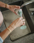 Wearing gold jewelry, a person uses the mushie Silicone Baby Bottle Brush to clean a green glass under running water in a kitchen sink, as water fills the basin below.