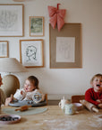 A woman and two young children smile and laugh at a table covered with baking supplies, brightened by Bluey x Mushie Silicone Placemats from mushie. Behind them, the wall features framed art and a big red bow.