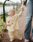 A baby in yellow checkered clothes, holding a mushie Unicorn Figurine Teether and a pacifier, walks by the water while grasping an adult's hand. The partly visible adult wears jeans and pink sneakers.