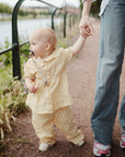 A small child in yellow checkered clothes with a mushie Linen Pacifier Clip 2-Pack holds an adult’s hand while walking on a gravel path by the water. The adult wears blue jeans and pink sneakers as greenery lines the path.