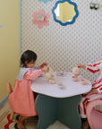 Two young children in colorful clothes play with the Mushie Silicone Tea Play Set at a small table in a pastel-themed, softly lit playroom decorated with playful wall art.