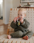 Toddler in green outfit on chair chews a mushie Teething Gift Set sensory teether.