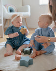 Two babies in blue play on a round mat with mushie Nesting Stars Toy as an adult assists. A crib and chair add to the softly lit, cozy Danish Hygge atmosphere—ideal for encouraging fine motor skill development.