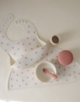 The mushie Silicone Placemat set in soft pink and white includes a bib, placemat, bowl with spoon, cup, and textured ball. The bib and placemat feature a pink bow pattern and are neatly arranged on a light surface.