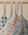 Four mushie Water Resistant Wet Bags with zippers hang by cream loops on a wooden rod, featuring green checks, bees, colorful flowers, and blue florals—perfect for diaper bag storage.