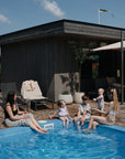 A woman and three children, one wrapped in a mushie Bear Robe, sit by a small blue pool splashing water on a wooden deck next to a modern house, while another woman relaxes nearby on a sunny day.