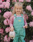 A smiling toddler in green overalls and a pink shirt stands before bright pink rhododendrons, holding a floral teapot, a pink flower, and a spill-proof mushie Snack Cup.