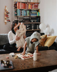 A woman on a couch holds a child in an organic cotton hooded towel by mushie, while another child wears the mushie Bear Poncho Towel and stands by a wooden table with food and a bottle in a cozy, book-filled living room with colorful cushions.