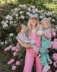 A woman in pink overalls and a blue cap holds two young girls outdoors by pink flowers; one girl, a toddler, is holding the mushie Snack Cup. Both girls wear colorful outfits and look happy and playful.